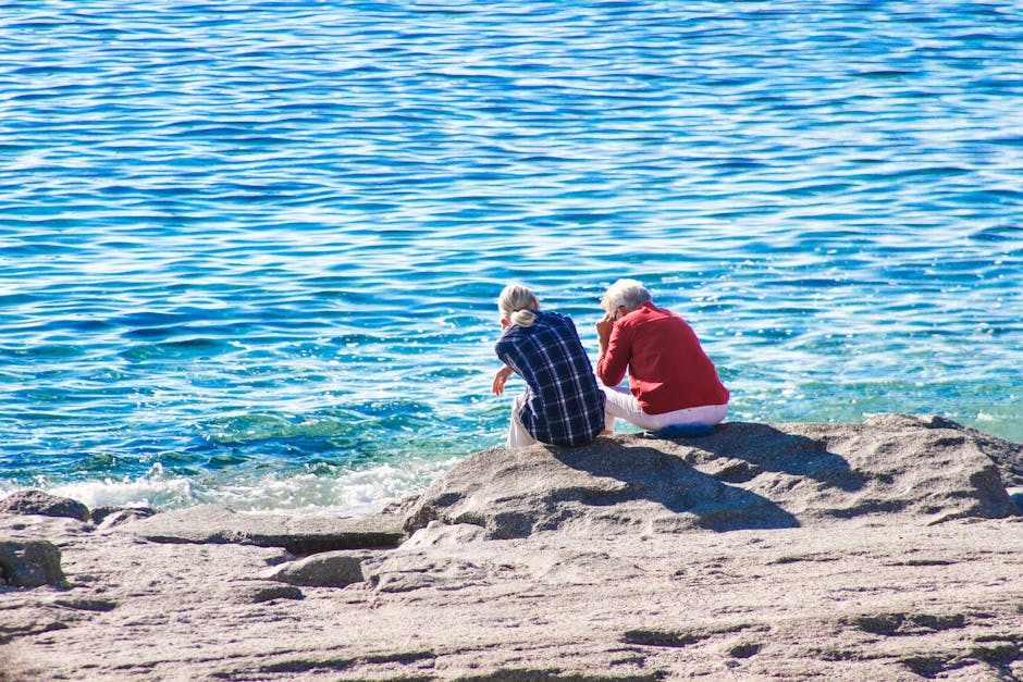 Two seniors sitting by the sea, enjoying a sunny day on the rocky shore.