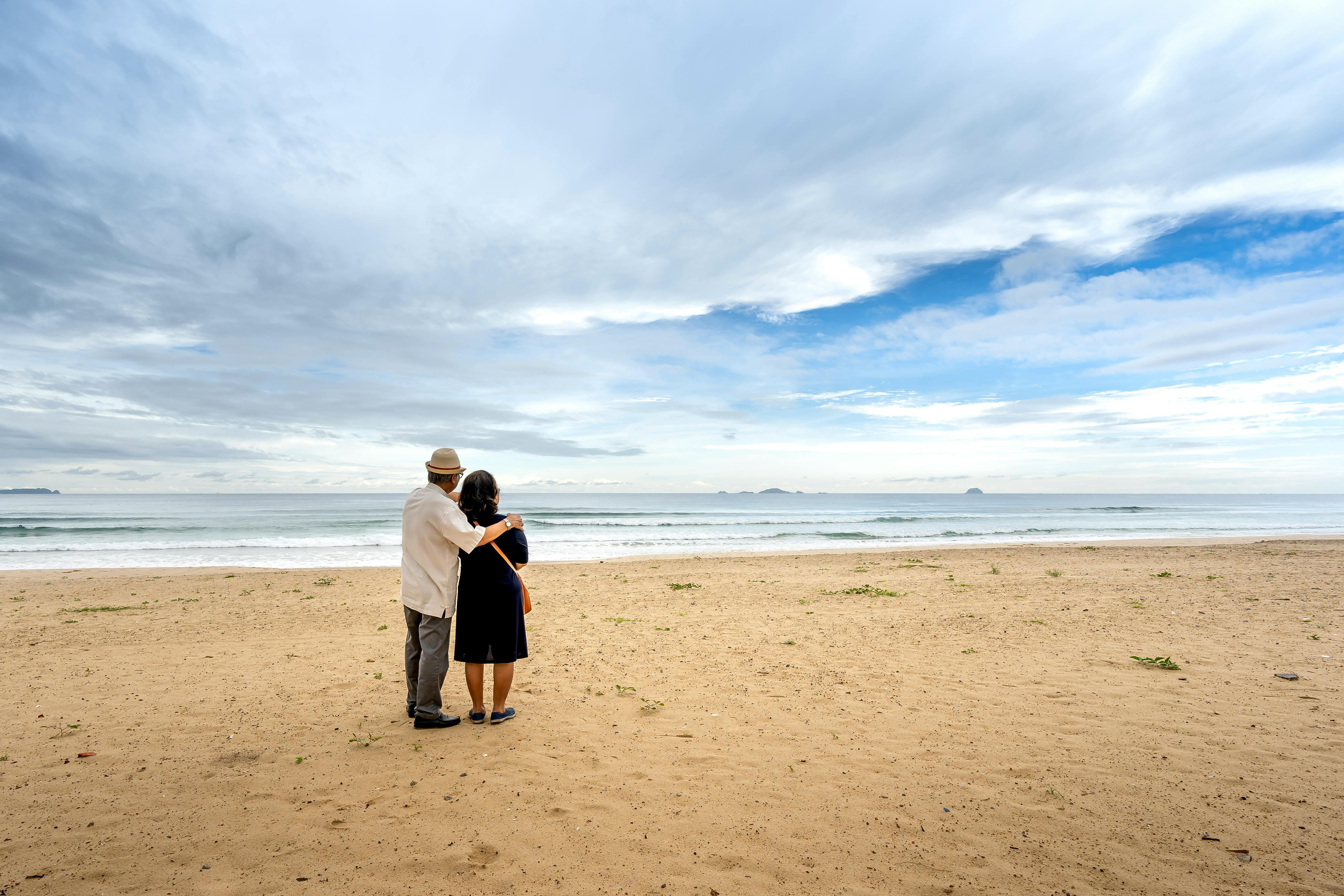 Couple standing on a wide quiet beach beneath a cloudy sky, looking toward the horizon — the long planning horizon of a 30-year retirement