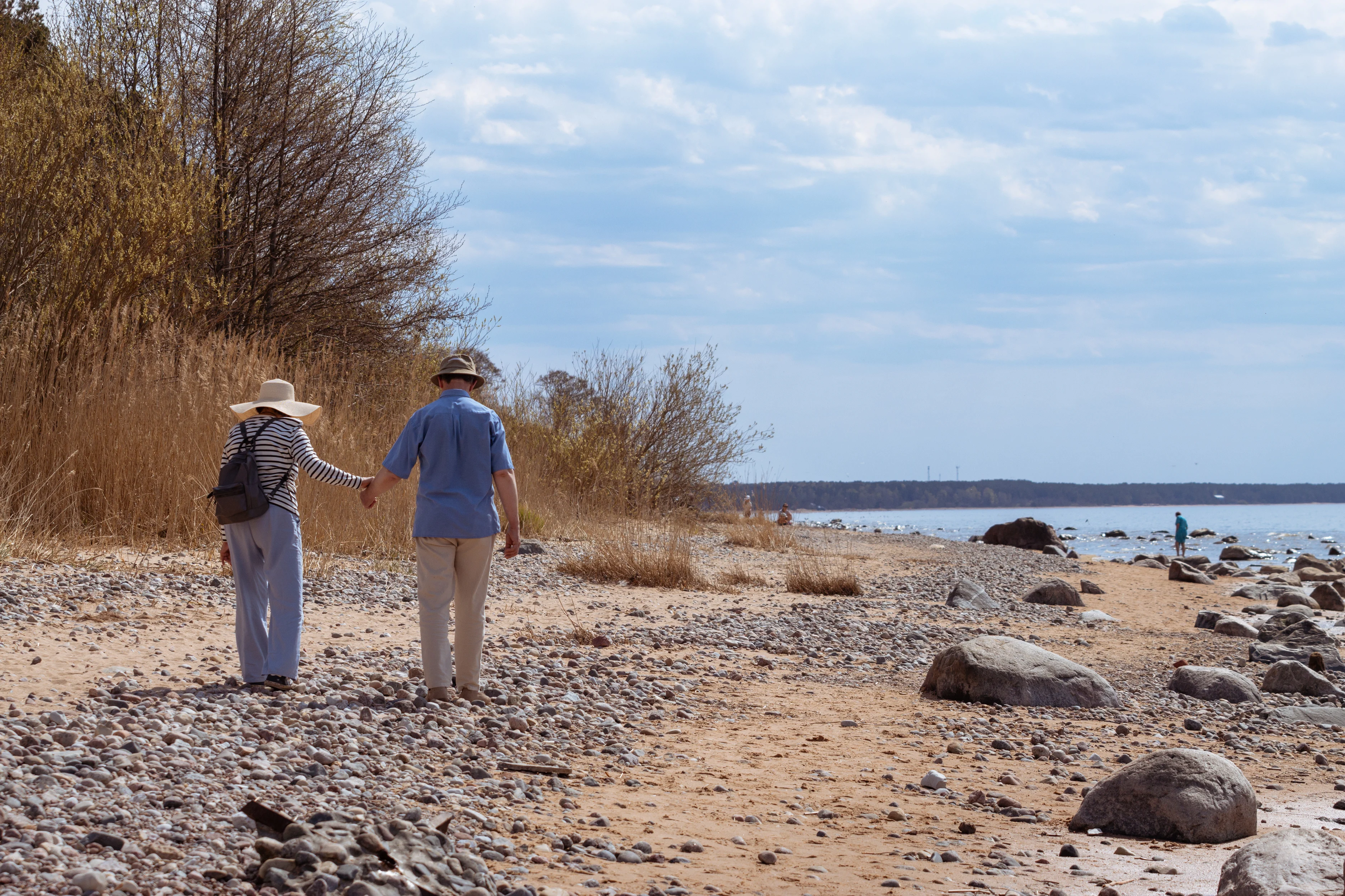 Couple in their early sixties walking hand-in-hand along a rocky autumn shoreline, symbolising the active early years of retirement at 60