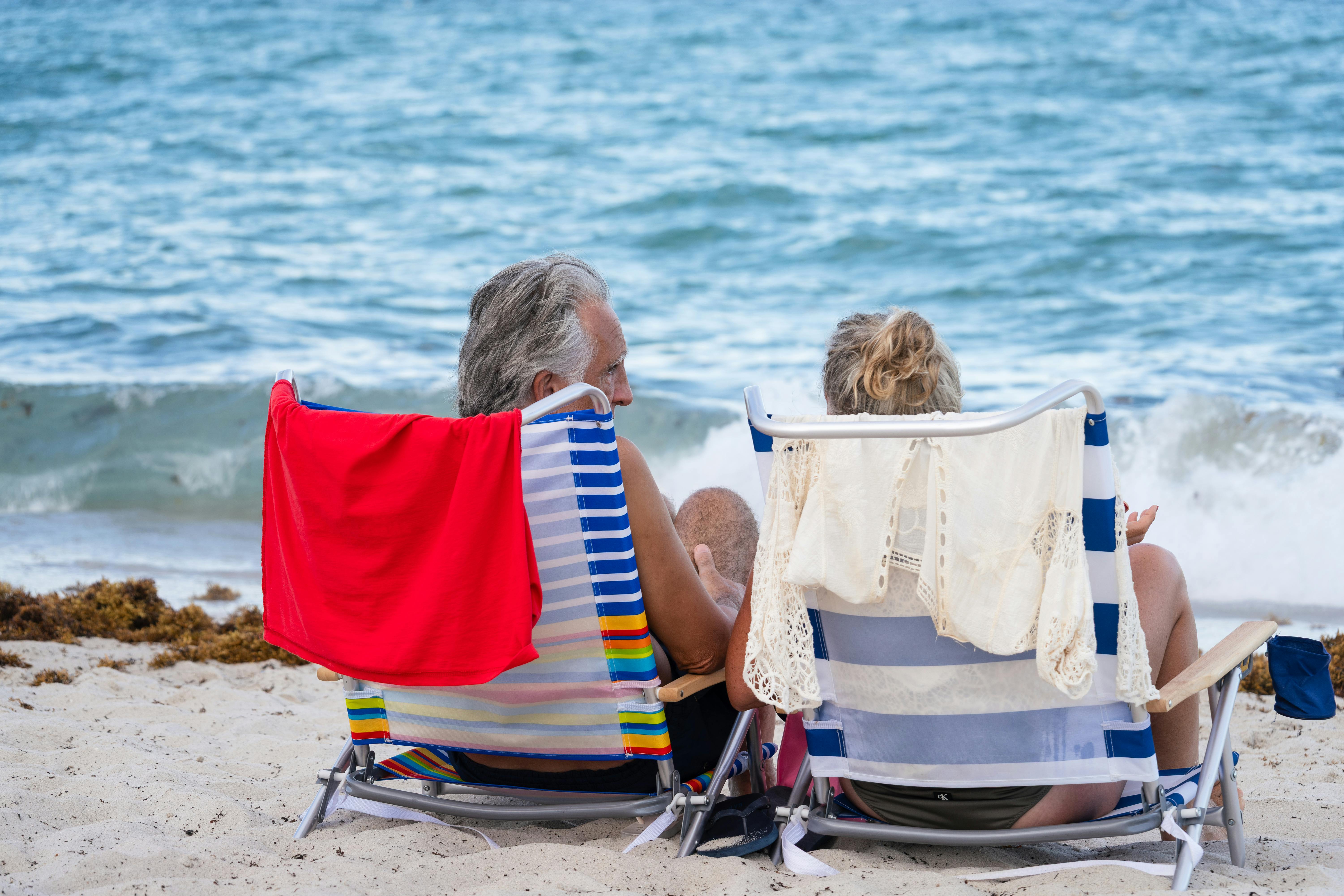 Mature couple seated in striped beach chairs watching the ocean, evoking the leisure time bought by retiring at 60