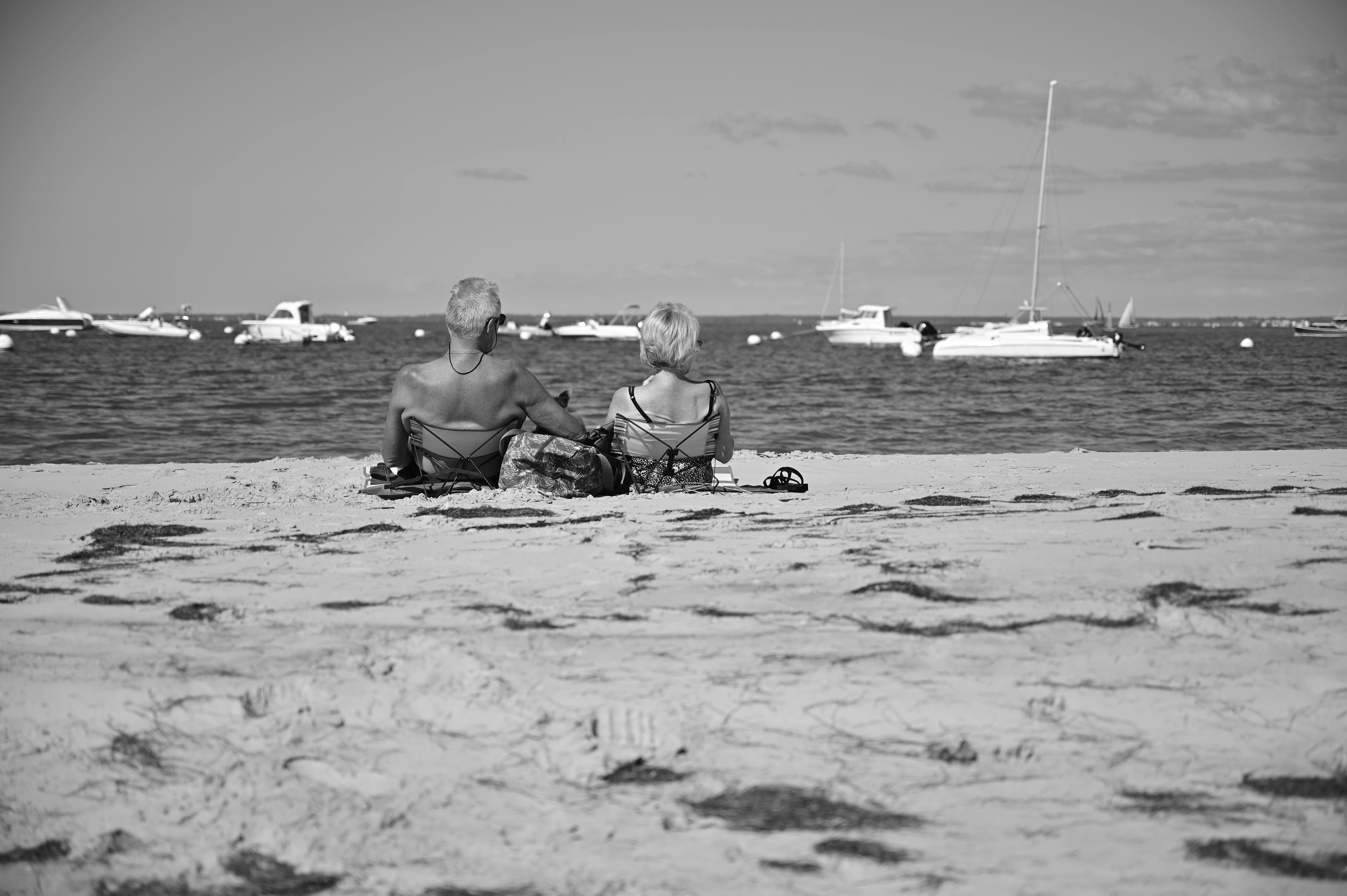 Black-and-white photograph of a couple sitting on a sandy beach watching distant sailboats — the long-term thinking the Social Security break-even math demands