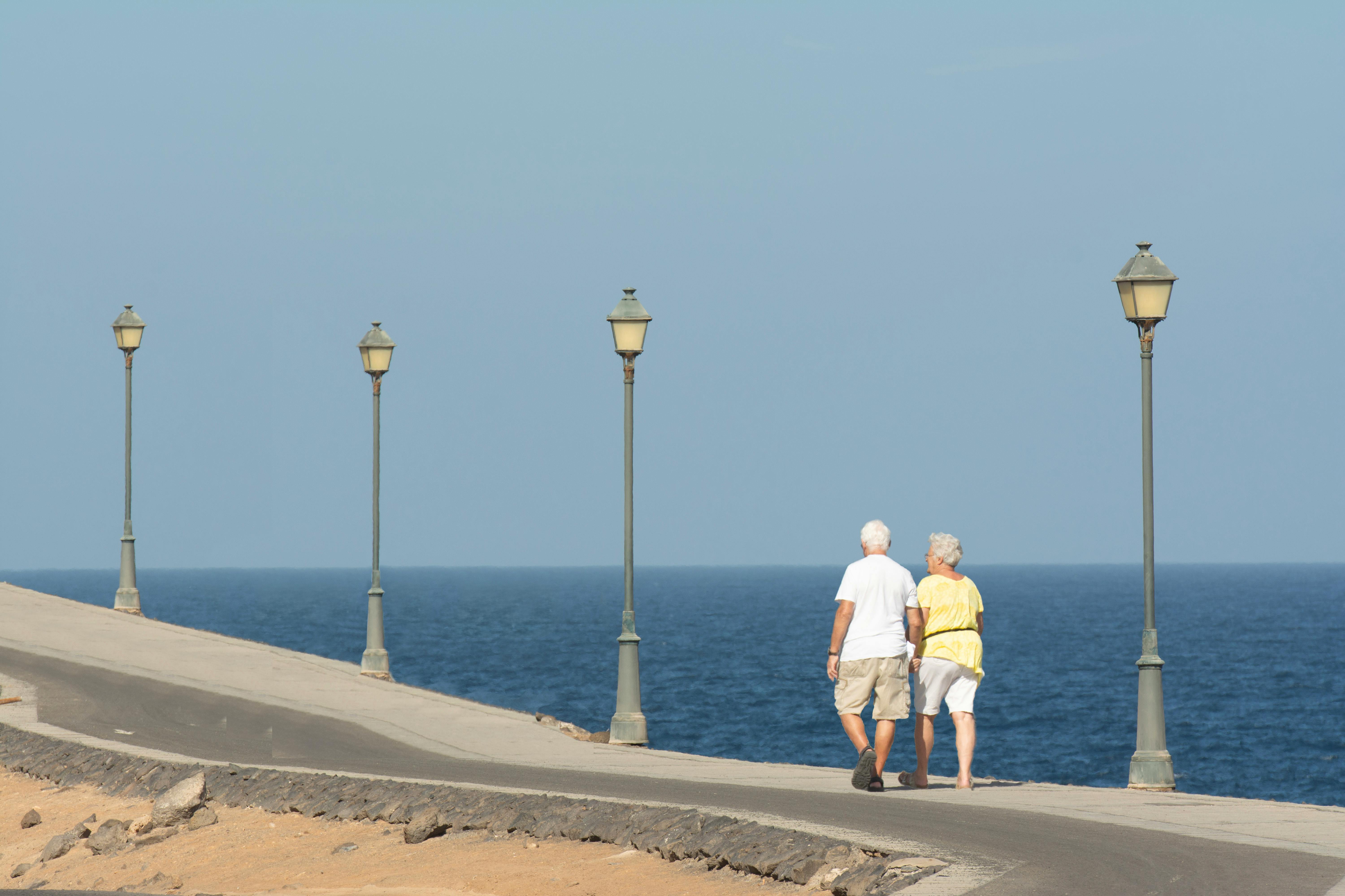 Retired couple walking together along a sunny seafront promenade past a row of lampposts and ocean — the steady 20-25 year horizon a retirement starting at 65 affords