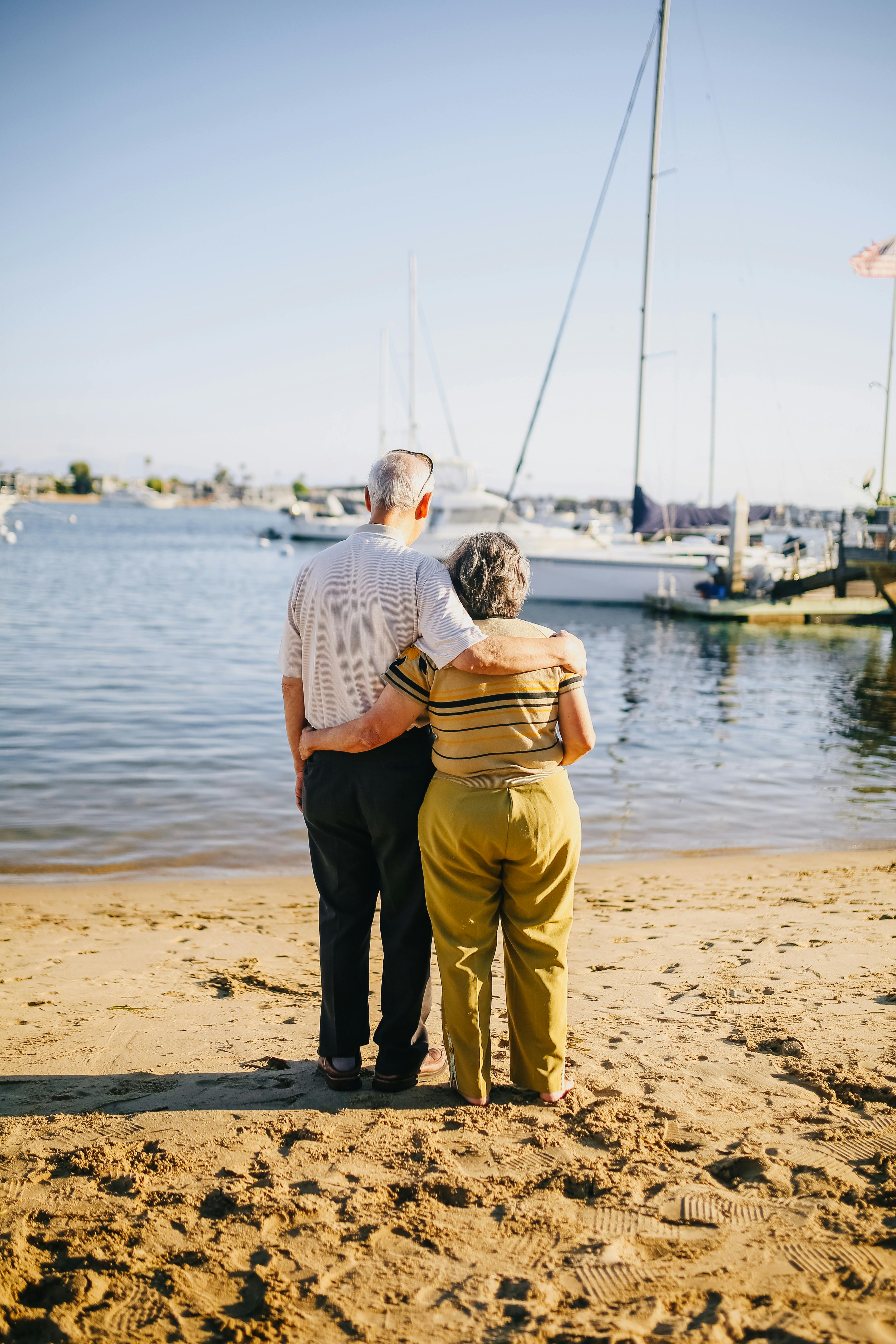 Retired couple embracing on a marina shore beside sailboats, symbolising the traditional retirement transition at age 65