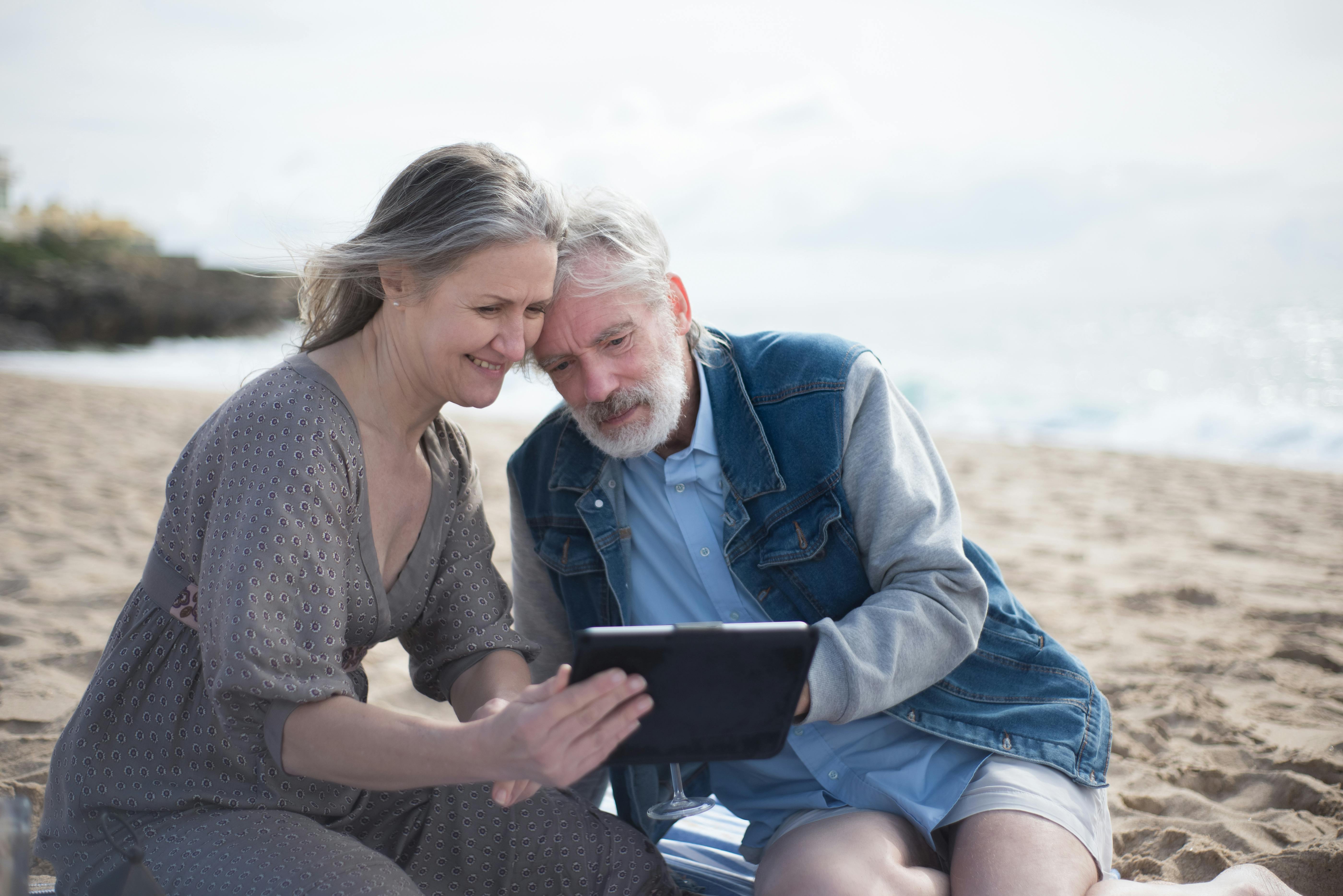 Older couple sitting close on a beach reviewing information together on a tablet — the work of comparing Medicare Parts A, B, C and D options