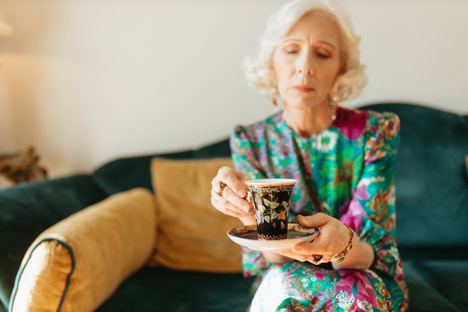 Senior woman with white hair savoring tea in a colorful dress on a cozy couch, adding a touch of elegance.